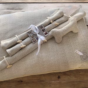 May include: A beige linen dog bed with a bone-shaped toy and three rolled-up linen toys tied with a white ribbon. The toys have small wooden buttons.