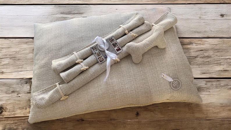May include: A beige linen dog bed with a bone-shaped toy and three rolled-up linen toys tied with a white ribbon. The toys have small wooden buttons.