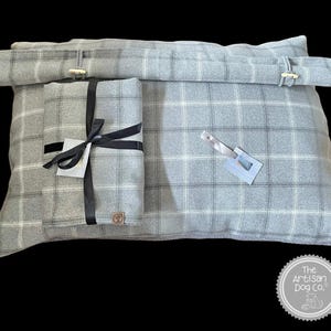 May include: A gray and white plaid dog bed with a removable, gray and white plaid pillow. The pillow is tied with a black ribbon and has a brown leather tag with the text "The Artisan Dog Co.".