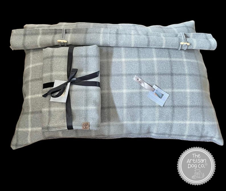May include: A gray and white plaid dog bed with a removable, gray and white plaid pillow. The pillow is tied with a black ribbon and has a brown leather tag with the text "The Artisan Dog Co.".