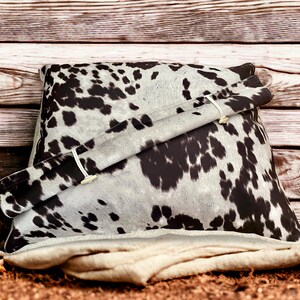 May include: A black and white cowhide patterned pillow with a white fabric flap and two buttons. The pillow is sitting on a rustic wooden background.