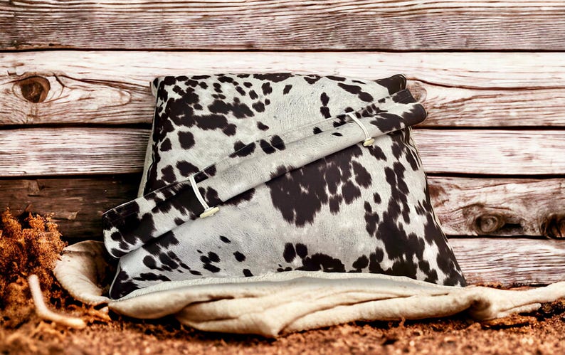 May include: A black and white cowhide patterned pillow with a white fabric flap and two buttons. The pillow is sitting on a rustic wooden background.