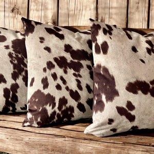 May include: Three brown and white cowhide patterned throw pillows on a wooden bench.