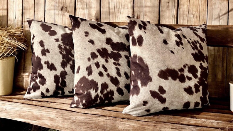 May include: Three brown and white cowhide patterned throw pillows on a wooden bench.