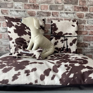 May include: A brown and white cowhide dog bed with a brown bone-shaped toy and a white stuffed dog toy. The bed has a black border.