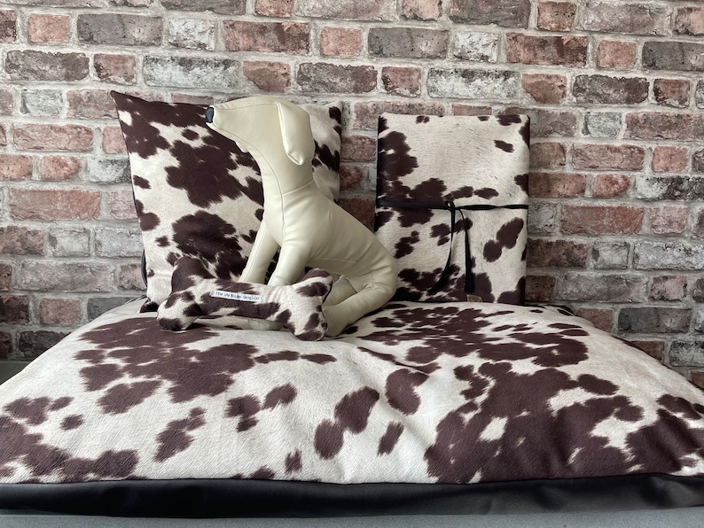 May include: A brown and white cowhide dog bed with a brown bone-shaped toy and a white stuffed dog toy. The bed has a black border.