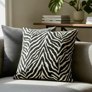 May include: A square cushion featuring a black and white zebra print. It sits on a grey sofa, next to a plain grey cushion. The room has a neutral colour scheme, with a plant and books visible in the background. The cushion measures approximately 40cm x 40cm.