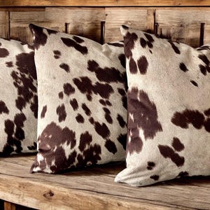 May include: Three brown and white cowhide patterned throw pillows on a wooden bench.