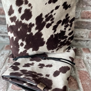 May include: Two brown and white cowhide patterned throw pillows with black leather trim. The pillows are stacked on top of each other. The pillow on top has a black leather tie.