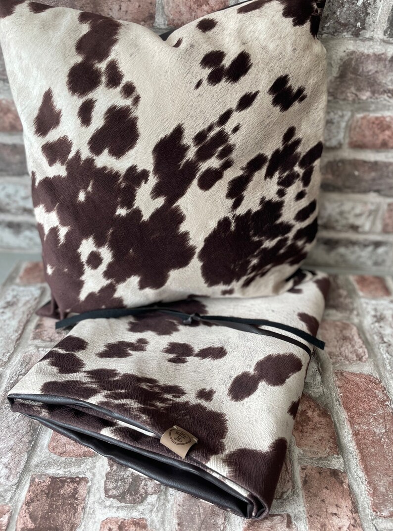 May include: Two brown and white cowhide patterned throw pillows with black leather trim. The pillows are stacked on top of each other. The pillow on top has a black leather tie.
