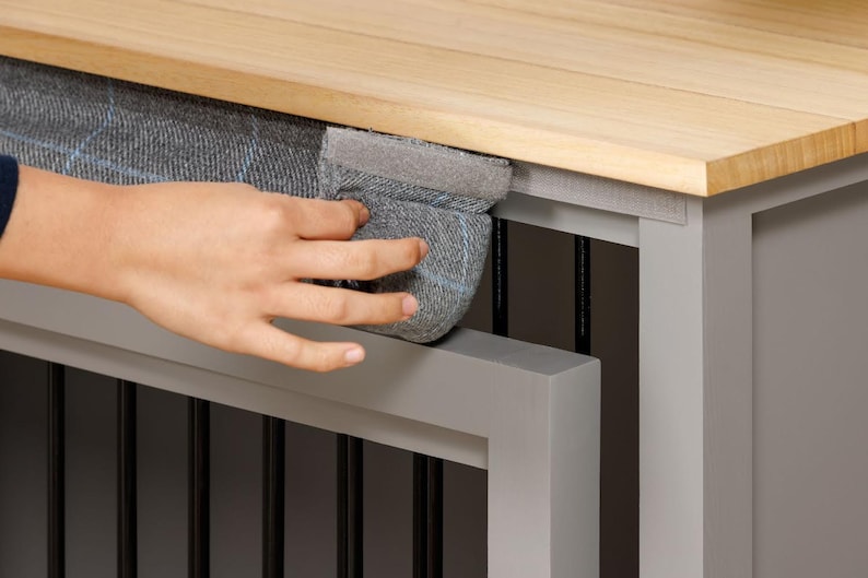 May include: A gray wooden dog crate with a gray fabric cushion on top. The cushion is secured with a gray velcro strap. The crate has a wooden top that can be lifted to access the inside.