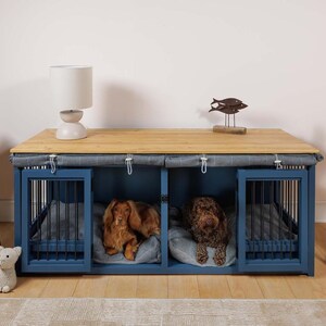 May include: A blue wooden dog crate with two compartments, each with a black metal door and a gray fabric cover. Two dogs are inside the crate, one brown and one black. The crate is sitting on a wooden table with a lamp and a decorative fish sculpture on top.