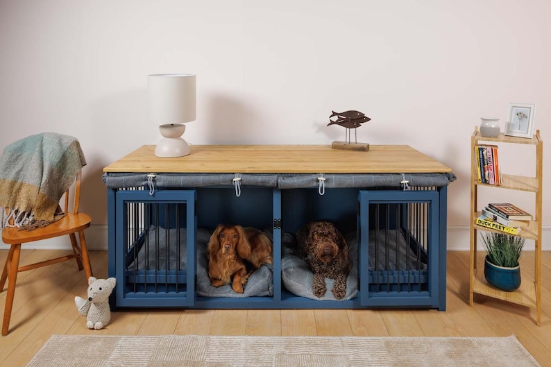 May include: A blue wooden dog crate with two compartments, each with a black metal door and a gray fabric cover. Two dogs are inside the crate, one brown and one black. The crate is sitting on a wooden table with a lamp and a decorative fish sculpture on top.