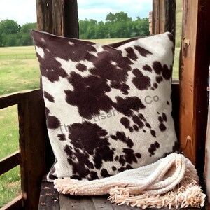 May include: A brown and white cowhide patterned throw pillow with a fringe throw blanket on a wooden surface. The Artisan Home Co. logo is visible in the image.