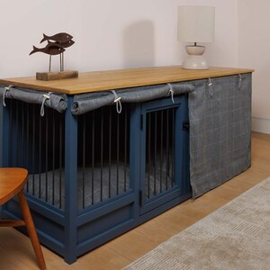 May include: A blue wooden dog crate with two compartments and a wood top. The crate has a fabric cover that can be pulled down to create a den-like space. The crate is sitting on a wooden floor with a rug in front of it.