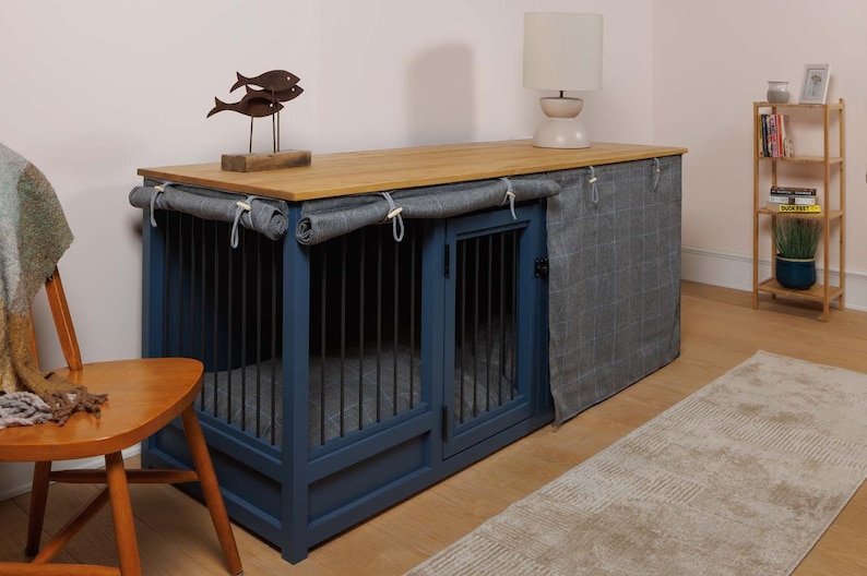 May include: A blue wooden dog crate with two compartments and a wood top. The crate has a fabric cover that can be pulled down to create a den-like space. The crate is sitting on a wooden floor with a rug in front of it.