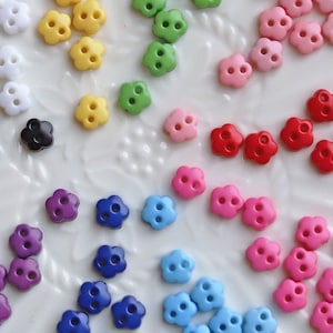 Puede incluir: Surtido de botones con forma de flor de colores, incluyendo blanco, negro, amarillo, verde, rosa, rojo, morado y azul. Los botones están dispuestos en un círculo sobre un plato blanco.