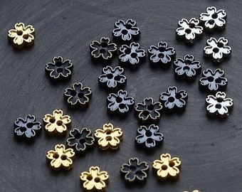 May include: A collection of small metal flower-shaped charms in gold, silver, and black colors. The charms are scattered on a black surface.