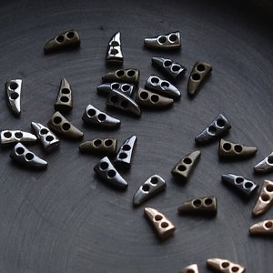 May include: A collection of small, metallic triangle-shaped buttons with two holes, scattered on a dark, round tray. The buttons vary in color, including silver, bronze, and a darker gunmetal. The tray has a slightly raised edge.