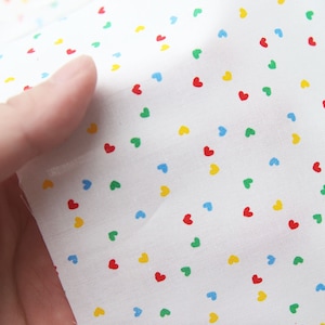 May include: White fabric with a repeating pattern of small, colourful hearts. The hearts are red, yellow, blue, and green. The fabric is folded in half and the text "#1" is visible in the bottom right corner.