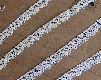 May include: White lace trim with a delicate scalloped edge. The trim is shown on a brown paper background.