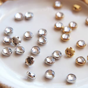 May include: A white ceramic dish with a gold ribbon bow on top. The dish contains a variety of small, round, clear rhinestones with silver and gold settings.