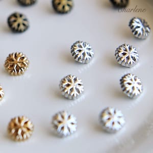 Könnte beinhalten: Eine Sammlung von silbernen und goldenen Knöpfen in Schneeflockenform. Die Knöpfe sind in einem Muster auf einer weißen Oberfläche angeordnet.
