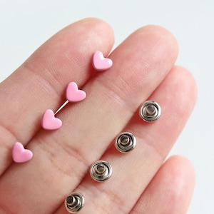 May include: A set of pink heart-shaped studs and silver metal snaps. The snaps are round and have a raised center. The hearts and snaps are held in a hand.