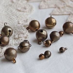 May include: A collection of small, gold-toned bells in various sizes and shapes. The bells are scattered on a white lace background.