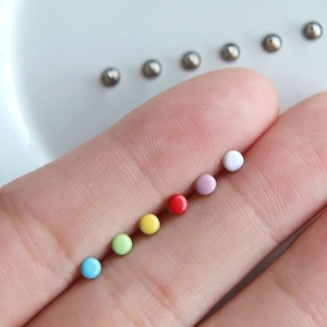 Może przedstawiać: Zestaw 7 małych, okrągłych, kolorowych koralików. Korale są ułożone w kolejności tęczy, zaczynając od niebieskiego, następnie zielonego, żółtego, pomarańczowego, czerwonego, różowego i białego.