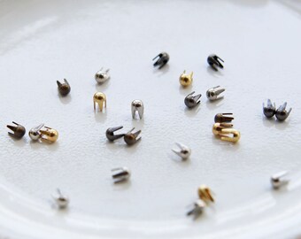 May include: Assortment of small metal rivets in silver, gold, and black colors on a white plate. The rivets have a rounded head and a pointed end.