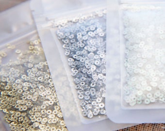 Könnte beinhalten: Drei Beutel mit Pailletten in Silber, Gold und Weiß. Die Pailletten sind klein und rund und in durchsichtigen Plastiktüten verpackt.