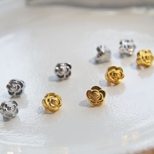 May include: Assortment of small rose-shaped charms in gold and silver tones. The charms are arranged on a white surface.