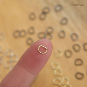 May include: A close-up of a gold-colored metal ring, held between a finger and thumb. The ring is a D-ring, with a flat, open top and a rounded bottom. The ring is surrounded by a scattering of similar rings, some gold and some silver.