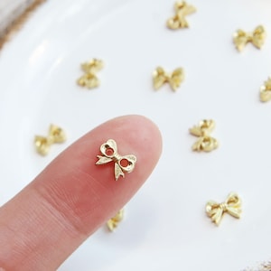 Könnte beinhalten: Nahaufnahme eines goldenen Metallbogens, der von einem Finger gehalten wird. Die Schleife ist klein und zart und hat eine leicht strukturierte Oberfläche. Im Hintergrund sind mehrere andere Schleifen auf einer weißen Oberfläche verstreut.