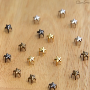 Może przedstawiać: Zestaw metalowych gwiazdkowych ćwieków w kolorze złotym, srebrnym i czarnym. Ćwieki są ułożone na drewnianej powierzchni.