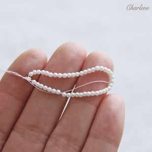 以下が含まれることがあります： 白い糸に白い真珠のビーズが通されたものを、手元で持っている様子のクローズアップ写真です。