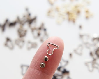 May include: Silver metal hook and loop fasteners, also known as bra strap adjusters, with a silver metal button. The fasteners are on a finger.