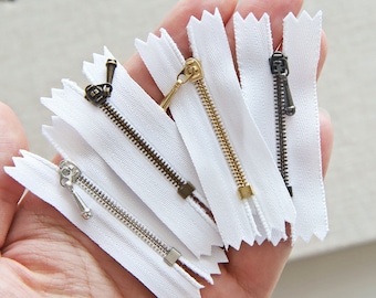May include: Five white zipper tapes with different colored metal zipper pulls. The zipper pulls are gold, silver, and bronze.