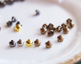 May include: Assortment of small metal star-shaped embellishments in gold, silver, and bronze colors. The embellishments are arranged on a white surface.