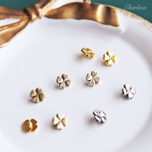 May include: A close-up of nine four-leaf clover charms in gold, silver, and bronze finishes. The charms are arranged on a white surface.