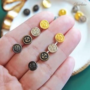 May include: A close-up of a hand holding a variety of small, round buttons with an anchor design. The buttons are in gold, silver, and black.