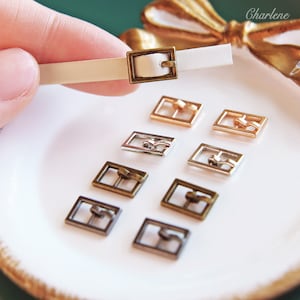 Könnte beinhalten: Sortiment von acht Metall-Gürtelschließen in verschiedenen Ausführungen, darunter Silber, Gold und Bronze. Die Schließen sind auf einer weißen Oberfläche angeordnet.