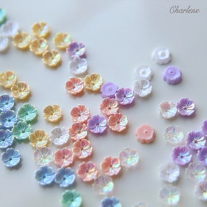 May include: Assortment of iridescent pastel flower-shaped beads in various colors, including pink, purple, blue, and yellow. The beads are scattered on a white surface.