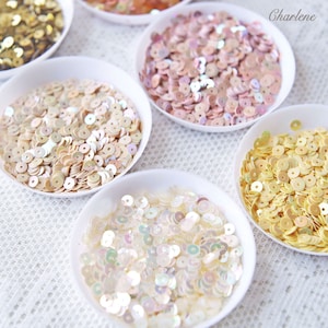 May include: Assortment of round sequins in various colours, including gold, pink, white, and beige, displayed in small white bowls.