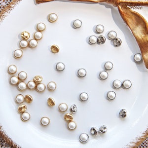 May include: A collection of white pearl buttons in various sizes and finishes, including gold and silver. The buttons are arranged on a white plate.