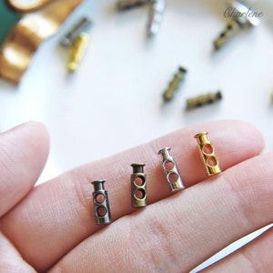 Może przedstawiać: Cztery metalowe stopery w różnych wykończeniach: srebrne, złote, antyczne mosiężne i czarne. Stopery mają dwa otwory i służą do regulacji długości sznurków lub pasków.