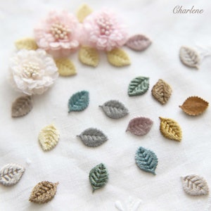 Può includere: Primo piano di un tessuto bianco con fiori e foglie ricamate in vari colori, tra cui rosa, giallo, verde, marrone e grigio. Le foglie sono disposte in uno schema sparso.