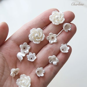 May include: A collection of white pearl flower-shaped beads. The beads are in various sizes and shapes, including single flowers and rosettes.