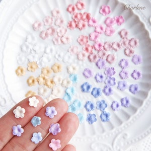 May include: An assortment of small, fabric flower embellishments in various pastel colours, including white, cream, light pink, lavender, and blue. The flowers are arranged in rows on a white plate, with a hand holding a selection of the flowers in the foreground.